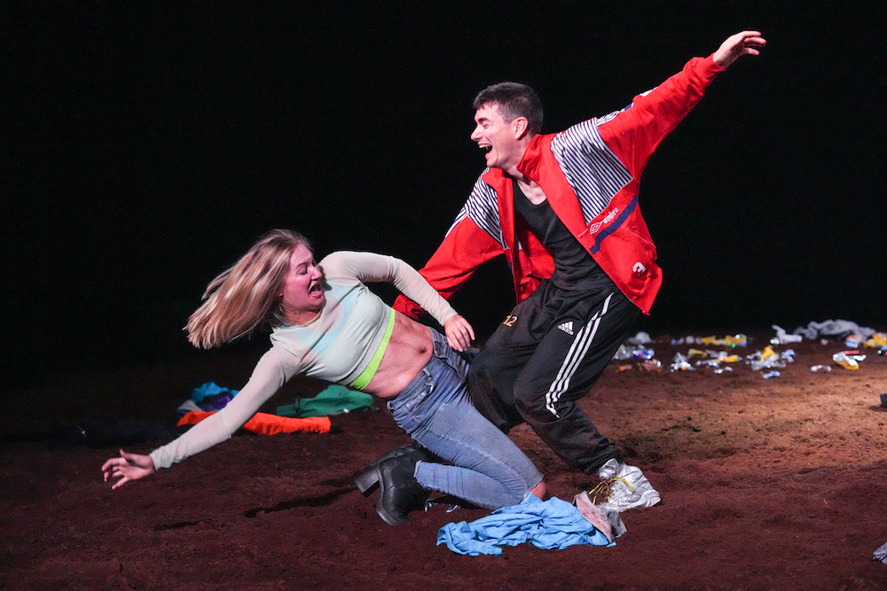 two people laughing so hard that one falls backwards and the other tries to pick her up. She is in a midriff top and jeans, he is in a red jacket wearing black and white sports pants.